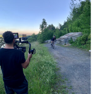Behind the scenes of a commercial shoot. Cameraman filming a woman biking on a rocky trail.