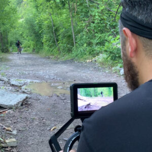 Behind the scenes of a commercial shoot. Cameraman filming someone biking through a wooded trail.