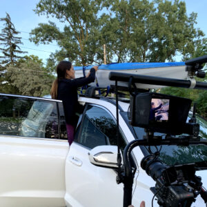 Behind the scenes of a commercial shoot. Camera is filming a woman strapping a kayak to her vehicle roof.