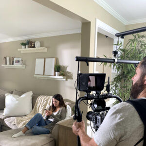 Behind the scenes of a commercial shoot. Cameraman is filming a woman on a couch on her phone.