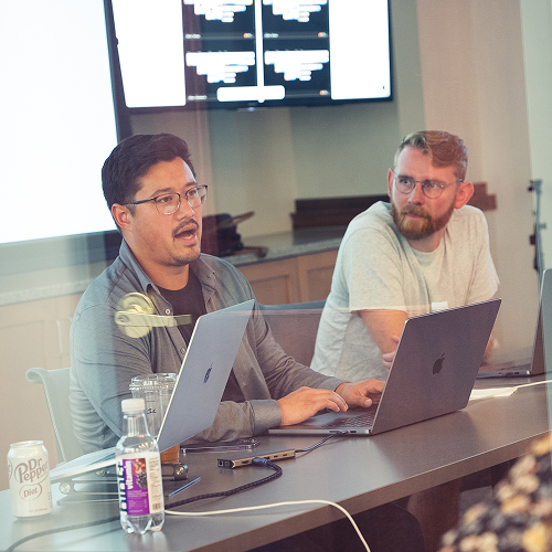 Two men in a conference room with laptops in front of them presenting something to the rest of the room