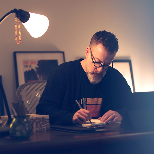 Man sitting at desk with a desk light on him as he is writing in a notebook
